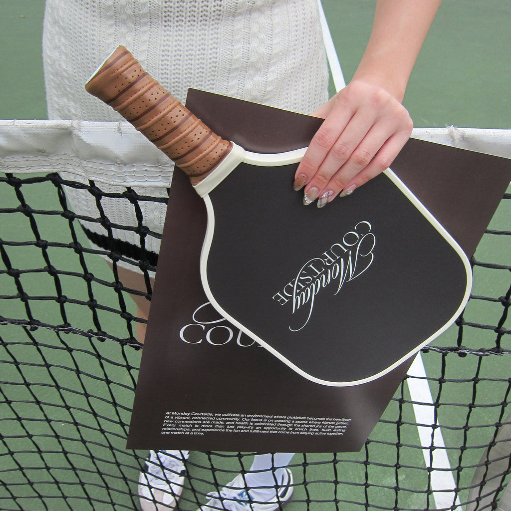 Person holding a brown pickleball paddle against a tennis court background.