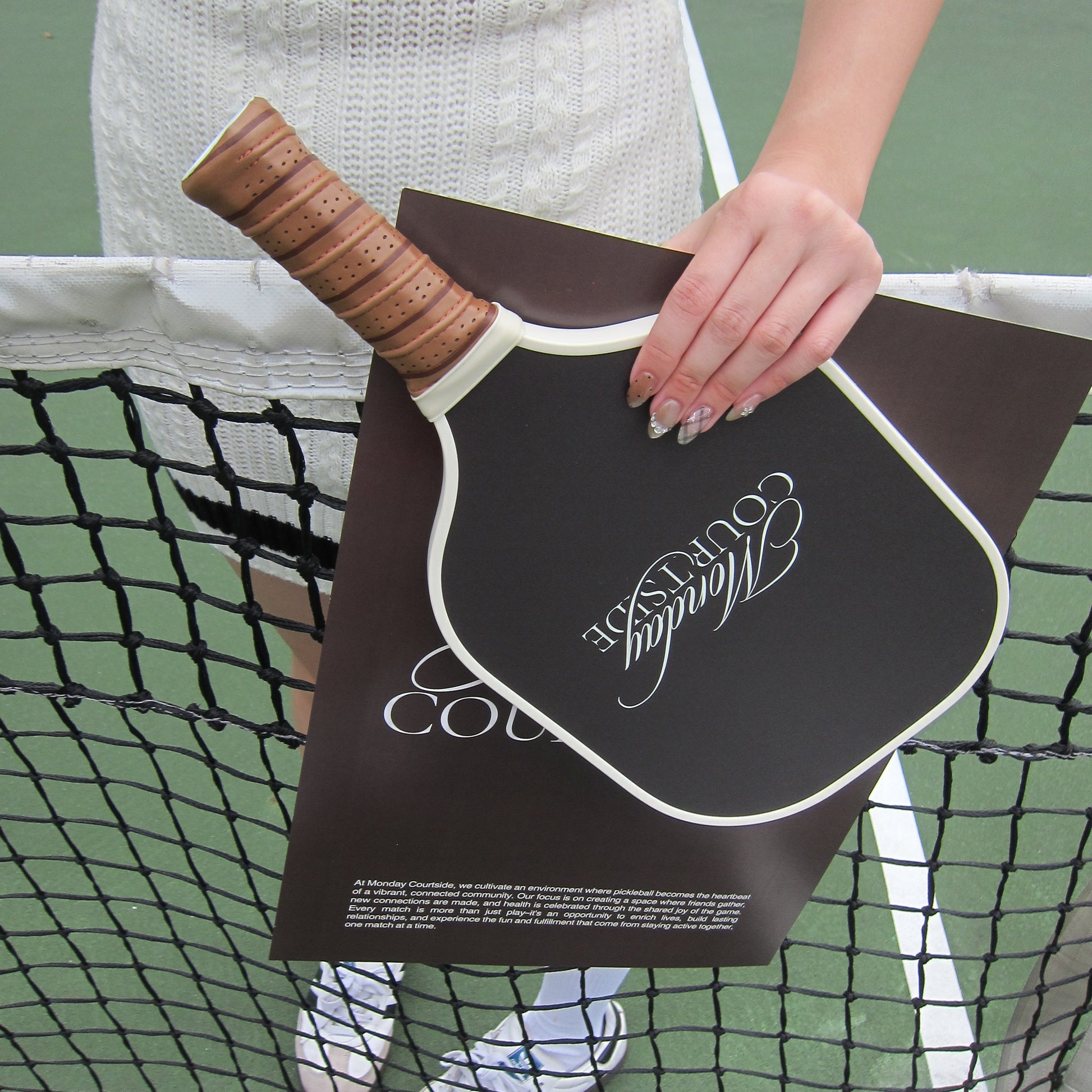 Person holding a brown pickleball paddle against a tennis court background.